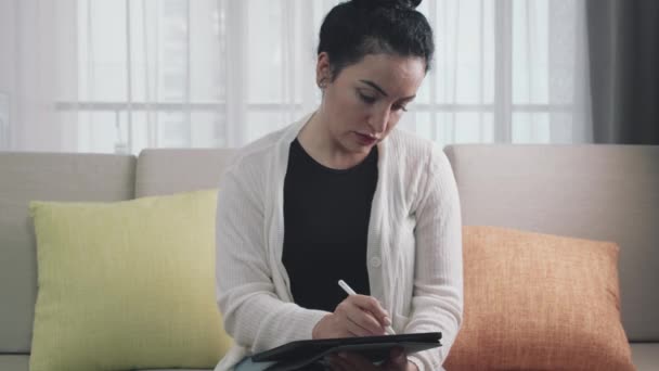 女性は自宅のソファに座って、デジタルタブレットコンピュータでペンを描き、書き込み、使用します。創造者として、何か新しい、専門家の創造者として、明るく明るいエネルギーの宣言 — ストック動画