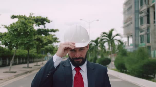 Portrett sjefsingeniør i dress, justerer sin hvite hjelm, ser rett inn i kameraet mot et bakteppe av trær og en byggeplass i en helt ny bydel – stockvideo
