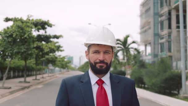 Portret starszego inżyniera przemysłowego w garniturze biznesowym, biały kask bezpieczeństwa, patrząc bezpośrednio w kamerę na tle drzew i placu budowy nowego kwartału w mieście — Wideo stockowe