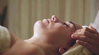 Kafasında havlu olan genç bir kadın, lüks bir sağlık merkezinde mükemmel cilt için yüz masajı ve spa tedavisi görüyor..