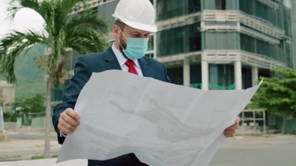 Mérnök vezető férfi fehér biztonsági sisakban, ász védőmaszkot visel, kezében az épület tervrajzával, ami az építkezésen kívül hat a Corona vírus közepette COVID 19 járvány vagy világjárvány. — Stock videók