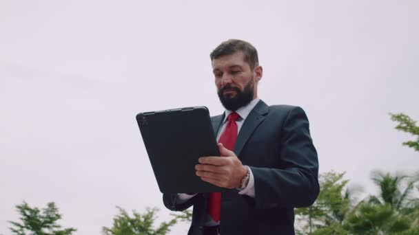 Porträtt av en seriös affärsman, skäggig affärsman, i en dyr kostym i röd slips, arbetar på en surfplatta dator i parken. — Stockvideo
