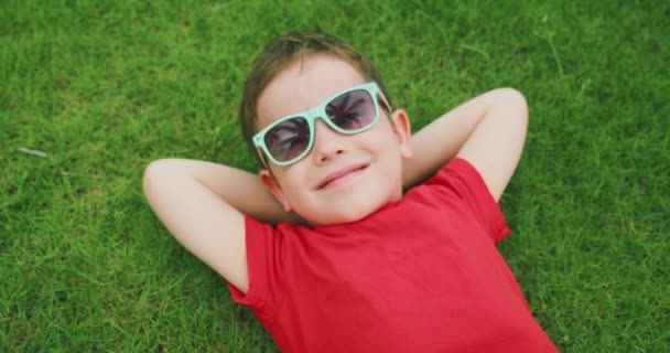 Ευτυχισμένο χαριτωμένο παιδί βρίσκεται στο πράσινο γρασίδι, ένα μικρό αγόρι με γυαλιά ηλίου βρίσκεται στο γρασίδι αναπαύεται. Ευτυχισμένη παιδική ηλικία. — Αρχείο Βίντεο