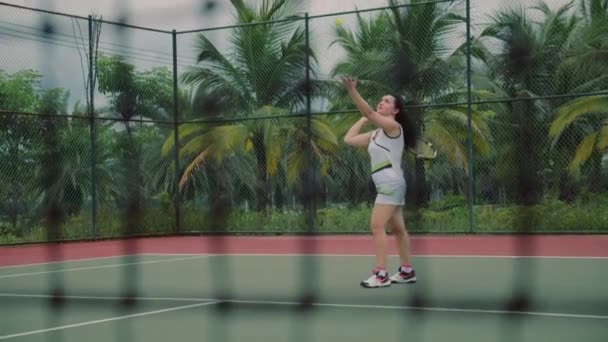 Vista lateral en cámara lenta de un joven atleta entrena el servicio de la pelota de tenis. Un aspirante a atleta adolescente juega tenis en la cancha. Chica activa golpeando la pelota duro mientras practica deportes — Vídeos de Stock