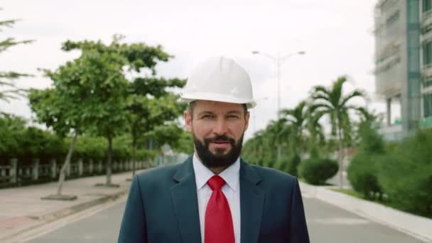 Portret starszego inżyniera przemysłowego w garniturze biznesowym, biały kask bezpieczeństwa, patrząc bezpośrednio w kamerę na tle drzew i placu budowy nowego kwartału w mieście — Wideo stockowe