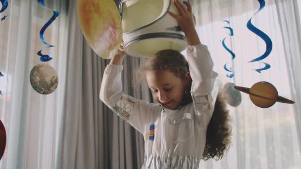 Child a girl puts on an astronauts helmet and plays in the space of her childrens room, the interior is hung with planets. Fantasy child astronaut plays with suspended space planets. — Stock Video