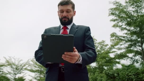 Ciddi bir iş adamının portresi, sakallı bir iş adamı, kırmızı kravatlı pahalı bir takım elbiseli, parkta bir tablet bilgisayar üzerinde çalışıyor.. — Stok video