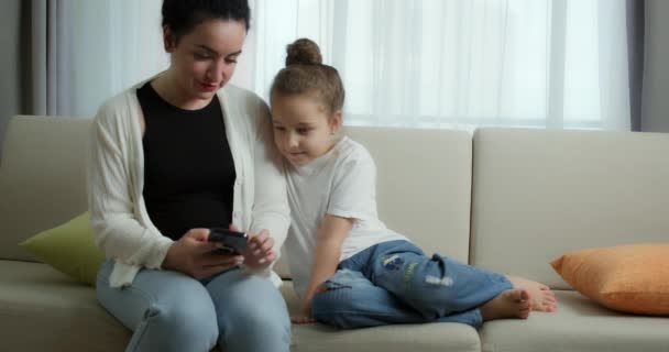 Happy mam en leuke dochter klein kind, moeder spelen met kind thuis ontspannen met een smartphone knuffelen zitten op de bank dochter, kijken grappige video 's, plezier hebben, genieten van het gezinsleven momenten — Stockvideo