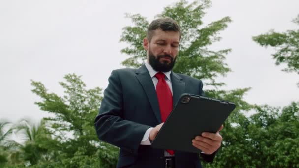 Portrait d'un homme d'affaires sérieux, homme d'affaires barbu, en costume coûteux en cravate rouge, travaillant sur une tablette à l'extérieur. — Video