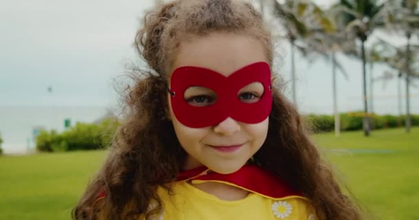 Porträt eines süßen lustigen Mädchens im Superheldenkostüm, in rotem Mantel und roter Maske, das einen Superhelden im Park spielt. — Stockvideo