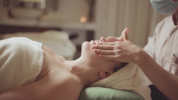 Giovane bella donna caucasica sdraiata sul letto spa ottenere il trattamento di massaggio facciale con aroma olio essenziale cura della pelle dal massaggiatore al salone di bellezza. Benessere cura della salute massaggio corpo spa concetto. — Video Stock