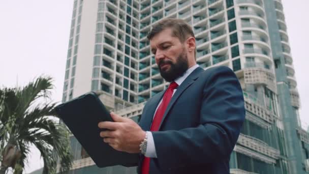 Retrato de un hombre de negocios serio, hombre de negocios barbudo, en un traje caro con una corbata roja, trabajando en una tableta en el parque. — Vídeo de stock