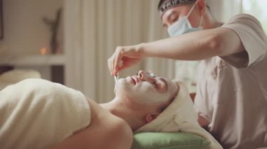 Spa yatağında uzanıp yüz masajı yapan rahat bir kadın lüks spa merkezindeki masör, spa merkezinde yüz maskesi takan bir kız. Sağlık, stres atma ve gençleştirme kavramı.