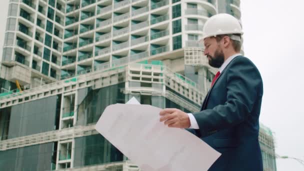 Ingeniero, varón en un desarrollador de casco de seguridad blanco en un sitio de construcción comprueba los dibujos en qué etapa de la construcción de edificios de varios pisos. — Vídeos de Stock