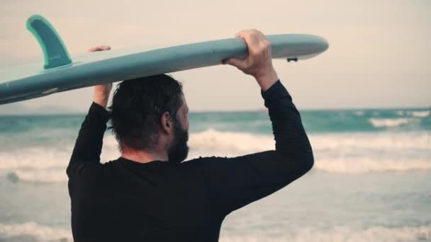 Senior Athlet im Alter geht mit einem Surfbrett an den Strand. Ein pensionierter Sportler fährt aufs Meer, reitet auf einem Surfbrett auf den Wellen. — Stockvideo