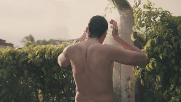 An athlete surfer takes a rain shower after kitesurfing. Shower in a private villa. Rear view of a man in swimsuit takes rain shower, tropical greenery on background. Guy washes in a private villa. — Stock Video