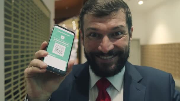 Retrato de cerca de un hombre de negocios mayor barbudo feliz que sonríe bonito y sostiene un teléfono con una aplicación que muestra el código verde dos vacunas de covid 19 y mirando a la cámara — Vídeos de Stock