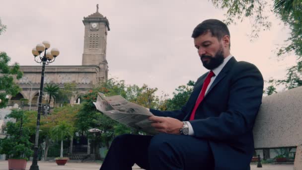 Νίκαιας ανώτερος επιχειρηματίας σε ένα ακριβό κοστούμι με μια όμορφη γενειάδα σε μπλε σακάκι κάθεται στον πάγκο στο πάρκο και διαβάζει μια εφημερίδα.Ώριμος επιχειρηματίας διαβάζει μια εφημερίδα κάθεται στο πάρκο στον πάγκο — Αρχείο Βίντεο