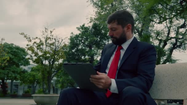 Un hombre barbudo serio y elegante con un traje caro lee un periódico en una tableta mientras está sentado en un banco en un parque al aire libre. Empresario con periódico y Tablet PC bebiendo café. — Vídeo de stock