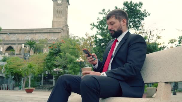Schwerer bärtiger Mann im teuren Anzug tippt etwas auf seinem Handy, während er draußen auf einer Parkbank sitzt — Stockvideo