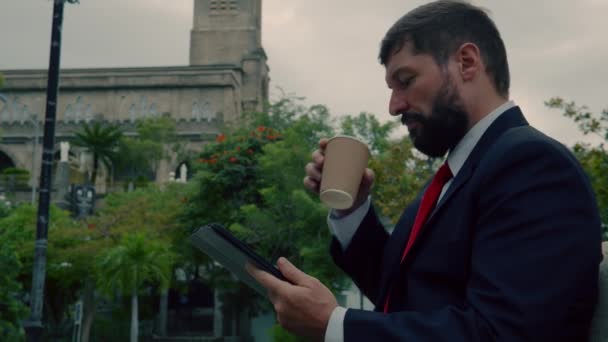 男子优雅的胡子工作在石碑上，坐在公园的长椅上。大胡子男子正在平板电脑上打字，喝咖啡. — 图库视频影像
