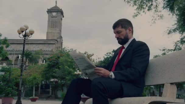 Empresario senior en un traje caro con una hermosa barba en chaqueta gris se sienta en el banco en el parque y lee un periódico. — Vídeo de stock