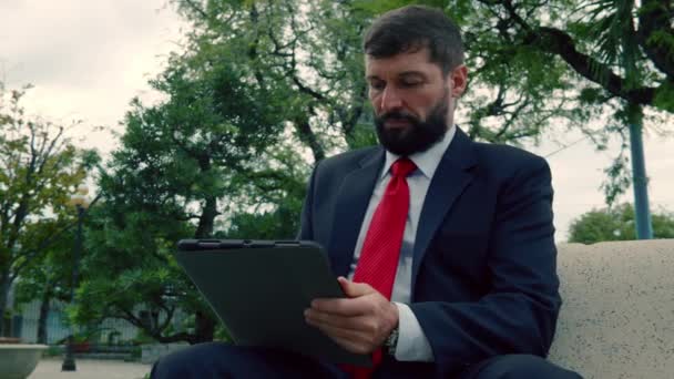 Homme d'affaires mature lisant un journal assis dans le parc sur le banc. Hommes d'affaires avec journal et tablette assis sur un banc dans un parc à l'extérieur. — Video