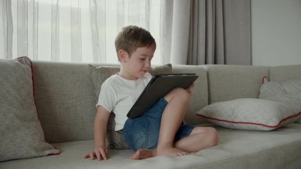 Aranyos óvodás fiú segítségével digitális tablet technológia eszköz online oktatás ül a kanapén otthon. Gyermek tech függőség koncepció. Szerkentyűs gyermek. — Stock videók