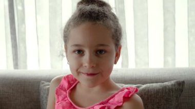 Portre Komik küçük kız kameraya bakıyor evde kanepede oturuyor, güzel yüzlü şirin çocuk kanepede oturuyor. Mutlu bir çocukluk kavramı.