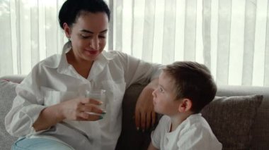 Tatlı anne, anaokulu çocuğuna bir bardak su verir, çocuk evdeki kanepede otururken cam bardaktan su içer. Ufaklık. Bir bardak su içen komik bir çocuğun portresi..