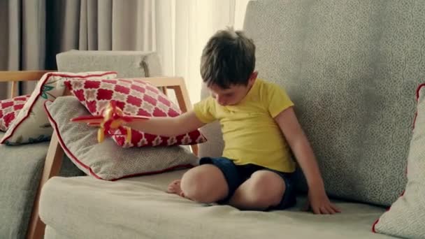 एक प्यारा छोटा प्रीस्कूलर लड़का सोफा पर घर पर एक खिलौना विमान के साथ खेल रहा है, सोफा पर बैठे एक प्यारा छोटा बेटा अपने हाथों में एक विमान लॉन्च करता है . — स्टॉक वीडियो