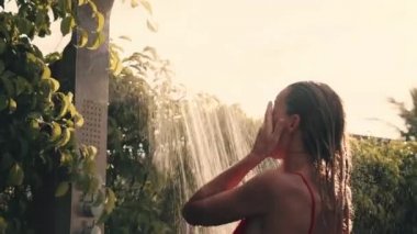 Genç sarışın kadın tropikal duş alıyor. Özel villada duş alıyor. Mayo giymiş bir kadın, arka planda tropik yeşillik ve yağmur banyosu yapar. Kafasını ve vücudunu yıkayan bir kız. Yavaş çekim