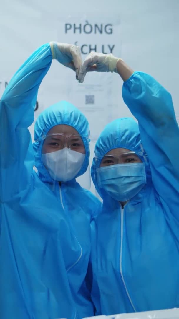 Två läkare i skyddsdräkt under epidemin visar tecken på liv och hjärta till stöd för andra som kan vara sjuka eller infekterade med coronavirus COVID 19. Vertikal video — Stockvideo