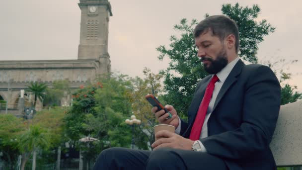 Komoly szakállas férfi drága öltönyben, aki gépel valamit a telefonján, miközben egy padon ül a parkban a szabadban. — Stock videók