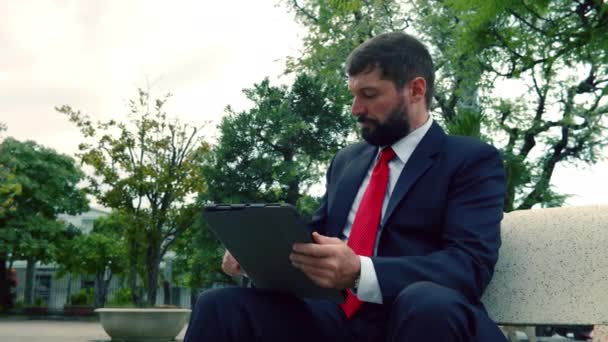 身穿昂贵西服的严肃而优雅的大胡子男人坐在公园的长椅上，一边在平板电脑上看报纸。拥有报纸和平板电脑喝咖啡的商人. — 图库视频影像