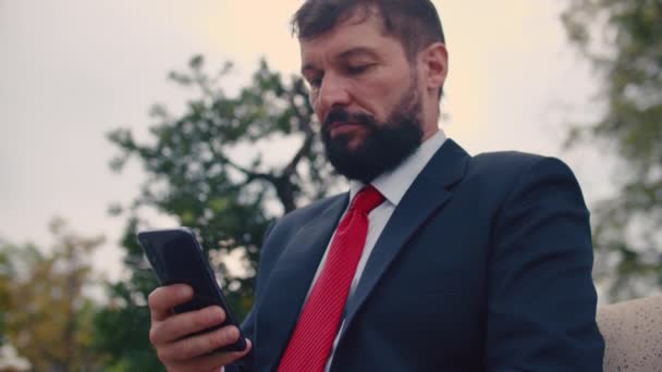Primer plano retrato hombre de negocios serio en un traje caro escribiendo algo en su teléfono mientras está sentado en un banco del parque al aire libre — Vídeo de stock