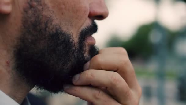 Um close-up de um queixo de homem, um homem idoso com uma barba acariciando sua barba, de pé na rua, olhando para o lado, contra um fundo turvo, o movimento de carros, close-up, câmera lenta. — Vídeo de Stock