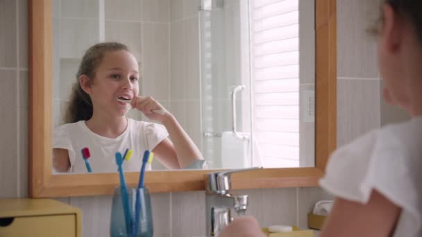 Portrait happy cute young teenage girl brushing teeth in bathroom and smiling. Children daily healthcare routine. Caucasian kid with white tooth looking at mirror isolated at home. Lifestyle. — Stock Video