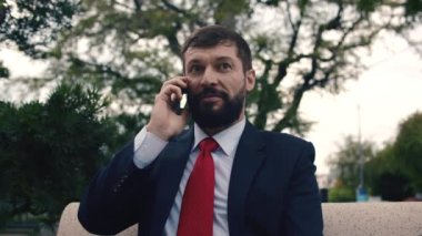 Parkta bir bankta otururken akıllı telefondan konuşan genç adam. Gözlüklü Yakışıklı Sakallı İş Adamı cep telefonu kullanıyor. İş için. Sosyal ağ. İnsanlar.