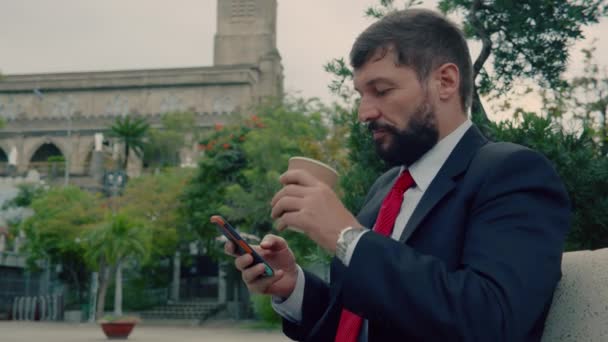 Geschäftsmann bärtiger Mann im teuren Anzug tippt etwas auf seinem Handy, während er draußen auf einer Parkbank sitzt — Stockvideo