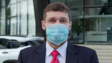 Coronavirus 'a karşı korumak için tıbbi maske takan yakışıklı iş adamı beyaz bir arabanın yanında duruyor ve arka planda binalar var ve kameraya bakıyor..