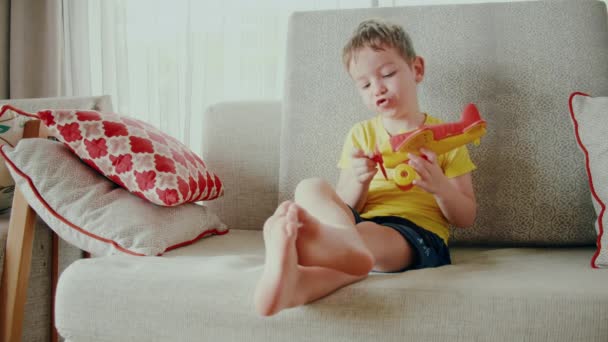 Evde kanepede oyuncak bir uçakla oynayan küçük tatlı bir anaokulu çocuğu kanepede oturan sevimli bir çocuk elinde bir uçak fırlatıyor.. — Stok video