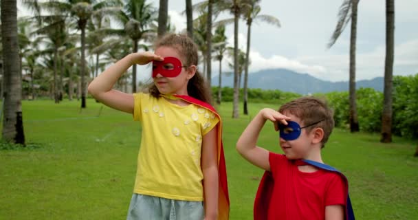 Przystojny chłopiec i dziewczyna przebrani za superbohatera, ubrani w niebiesko-czerwone płaszcze i superbohaterskie maski. Portret dzieci bohaterów przed tłem palmy, niebo i chmury rozejrzeć się w różnych kierunkach — Wideo stockowe