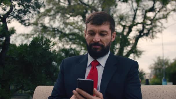 戴眼镜的英俊的胡子创业商人用手机。年轻人坐在公园的长椅上一边走路一边用智能手机说话。生意。社交网络。人. — 图库视频影像