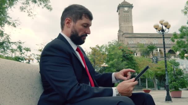 Hombre de negocios maduro leyendo un periódico sentado en el parque en el banco. Empresarios con periódico y tableta sentados en un banco en un parque al aire libre. — Vídeo de stock