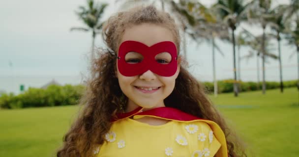 Portræt af en sød sjov barn pige i en superhelt kostume, i en rød kappe og en rød maske, spille en superhelt i parken. – Stock-video