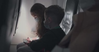 Koruyucu tıbbi maskeli bir uçakta küçük beyaz bir kız ve oğlan. Uçakta solunum cihazlı bir çocuk turist akıllı bir cep telefonuyla oynuyor. Coronavirus salgını covid-19 2019-ncov