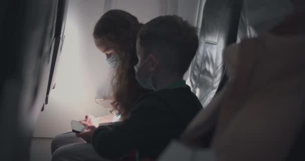 Klein blank meisje en jongen in een vliegtuig met beschermende medische maskers. Een kindertoerist in een vliegtuig met een ademhalingstoestel speelt op een smartphone. Coronavirus epidemie covid-19 2019-ncov — Stockvideo