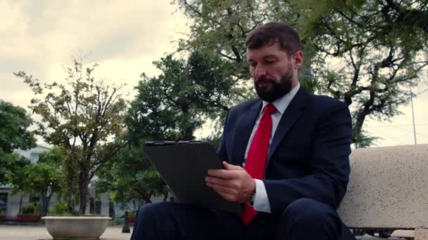 Un homme barbu élégant et sérieux en costume cher lit un journal sur une tablette alors qu'il est assis sur un banc dans un parc à l'extérieur. Homme d'affaires avec journal et tablette buvant du café. — Video