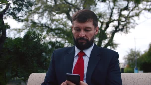 Komoly szakállas férfi drága öltönyben, aki gépel valamit a telefonján, miközben egy padon ül a parkban a szabadban. — Stock videók